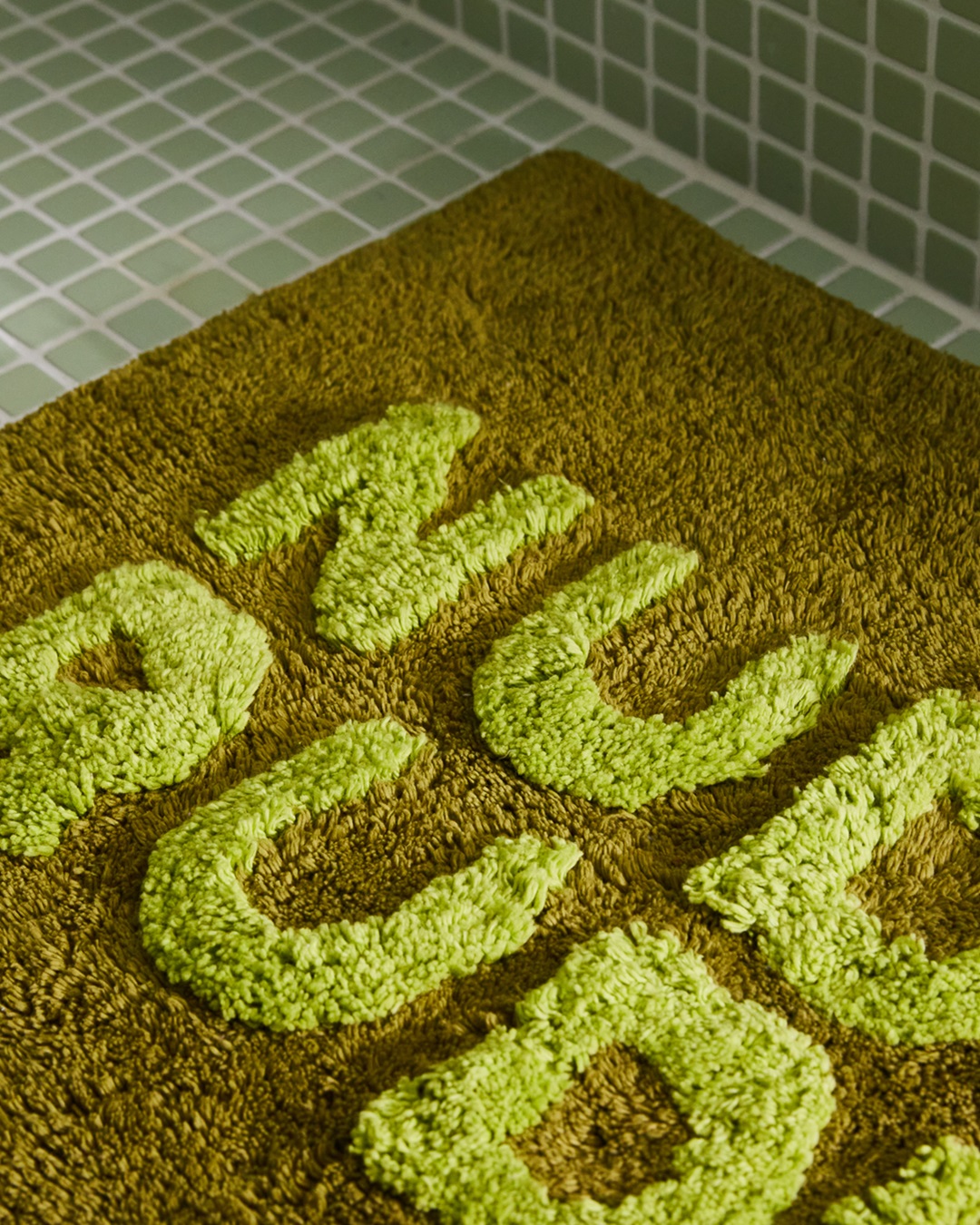 Green artichoke mini bath mat with Nudie Rudie on tiled bathroom floor